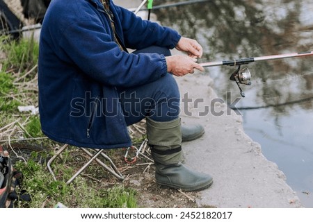 A man fisherman with a fishing rod in his hands sits on a chair in rubber boots on the river bank while fishing. Photography, hobby, lifestyle.