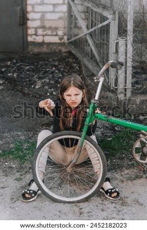 Little beautiful sad teenage girl, dissatisfied upset child showing dislike thumbs down sits near an old bicycle with a broken, punctured wheel tire outdoors. Photography, portrait.