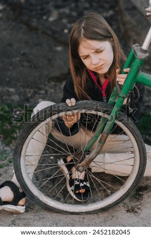 Little teenage girl, dissatisfied, distressed child sits near an old bicycle with a broken, punctured wheel tire outdoors. Photography, portrait. Royalty-Free Stock Photo #2452182045