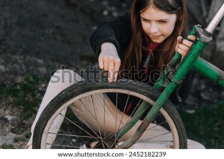 Little teenage girl, dissatisfied, distressed child sits near an old bicycle with a broken, punctured wheel tire outdoors. Photography, portrait. Royalty-Free Stock Photo #2452182239