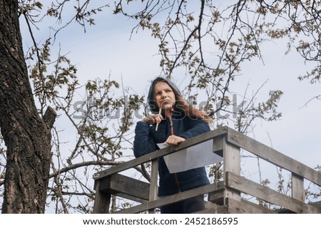 Beautiful pensive girl, inspired dreamy child teenager 8 years old stands on a wooden bridge outdoors in nature, writing poetry and drawing a picture of a landscape. Photography, portrait. Royalty-Free Stock Photo #2452186595
