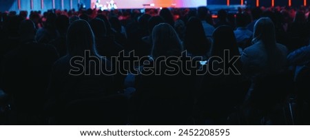 Audience viewing a presentation in a dark room with stage lights, banner size