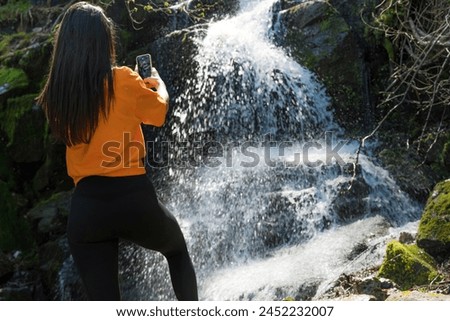 Woman standing in front of waterfall and taking pictures with the phone 