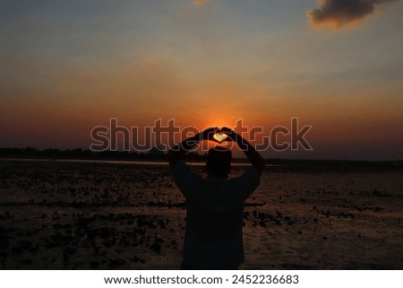 As the golden sunlight filters through the evening sky, creating a warm glow, two fingers form a heart shape, evoking a sense of romance and tranquility.
