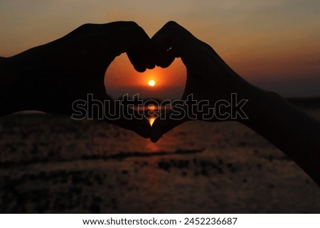 As the golden sunlight filters through the evening sky, creating a warm glow, two fingers form a heart shape, evoking a sense of romance and tranquility.