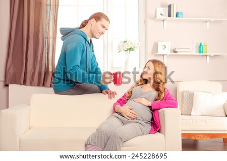 The husband holding cup of beverage for his pregnant wife while she is sitting on sofa at home. Happy pregnant family.