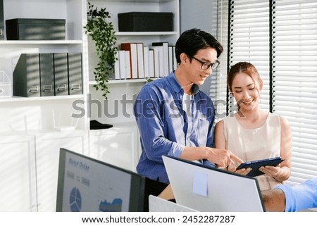 Casual Asian businessman and young female businesswoman discuss in the office, working on social media strategy, looking at a tablet with blur colleagues in the background. Image with copy space.