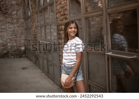 A beautiful girl with straw handbag, dressed denim shorts and a striped T-shirt goes on the street of the city, posing against the background of brick walls and large windows