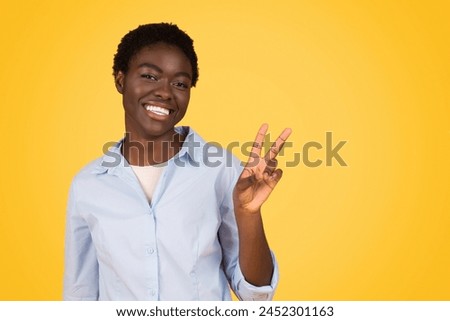 With a bright smile, this african american woman, a zoomer from generation z, makes a peace sign, isolated on a yellow