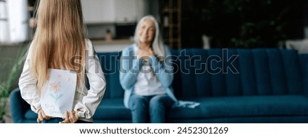 European small granddaughter holds behind back postcard with picture of happy senior lady in living room interior. Surprise birthday greeting and holiday celebration at home, copy space