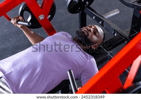 An African American young male athlete, a fitness model lying on a bench in a gym, lifting weights. He has short black hair, a beard, and is wearing a purple t-shirt.