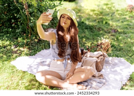 Young beautiful woman in trendy hat taking selfie on her phone on a green grass in a park