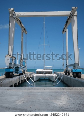 A large white travel lift is preparing its slings to lift a Lagoon 42 white catamaran yacht out of the Caribbean water.  Blue sky and horizon in the background. Royalty-Free Stock Photo #2452342641