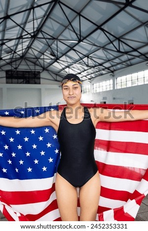 Biracial young female swimmer standing indoors wrapped in American flag, smiling at camera. She has a fit build, wearing a black swimsuit, cap, and goggles, unaltered. Royalty-Free Stock Photo #2452353331
