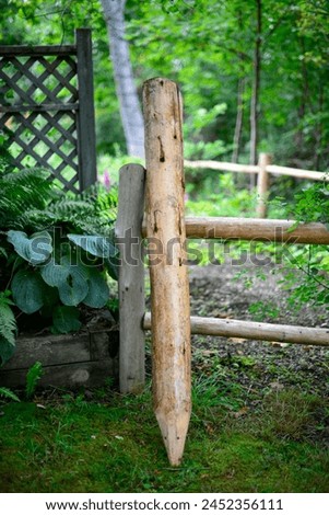 An aged wooden fence, adorned with rustic wooden stakes, encloses a rural farm pen. The lush green surroundings include trees, shrubs, and plants. Adjacent to the barrier stands a raised flower bed. Royalty-Free Stock Photo #2452356111