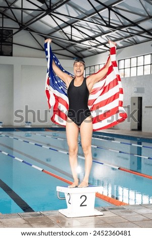 Young biracial female swimmer holding American flag, standing by pool indoors. She has dark hair, wearing goggles, and a black swimsuit, unaltered. Royalty-Free Stock Photo #2452360481