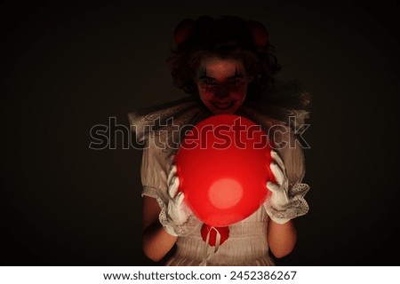 Scary Halloween. Portrait of a clown girl in a white circus outfit, who stands on a dark gloomy background and smiles, holding red balloon in her hand. Old circus concept. 