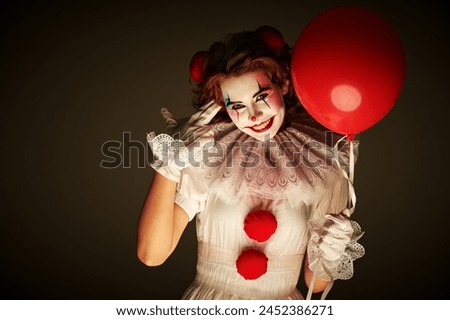 Old circus concept. Portrait of a clown girl in a white circus outfit, who stands on a dark gloomy background and smiles, holding red balloon in her hand. Scary Halloween. 