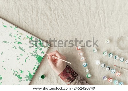 Painting by numbers with acrylic paints. Woman coloring picture by numbers, hand with brush and paints of different colors on linen tablecloth on table. Creative hobby, leisure activity at home.