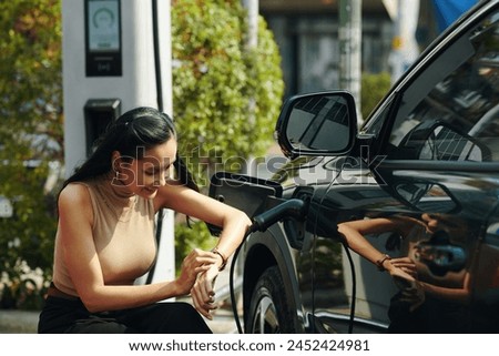 Smiling woman checking time on wristwatch when waiting for her electric car to fully charge Royalty-Free Stock Photo #2452424981