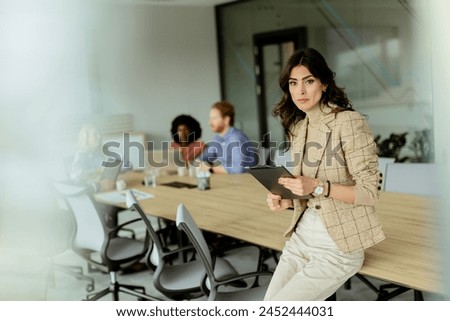 A poised woman with a tablet stands at the forefront of a collaborative office space, her team engaged in discussion behind her. Royalty-Free Stock Photo #2452444031