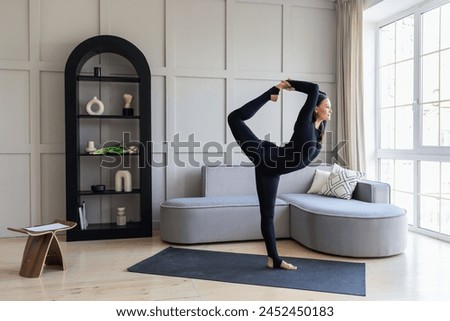 Yoga woman performing a variation of Natarajasana exercise, king of dance pose, training in black sportswear in the room standing on a mat, full length view, healthy lifestyle