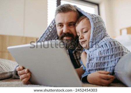 Little boy watching cartoon movie on tablet with father, lying under blanket on floor in kids room. Dad explaining technology to son, digital literacy for kids. Royalty-Free Stock Photo #2452454303