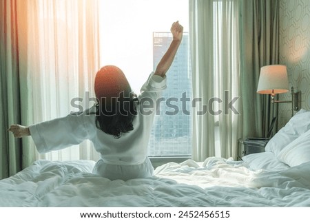 Hotel relaxation on lazy day with Asian woman waking up from good sleep on bed in weekend morning resting in comfort bedroom looking toward city view, having happy, work-life quality balance lifestyle Royalty-Free Stock Photo #2452456515