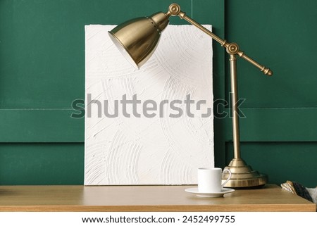 Minimalistic workplace with picture, lamp and cup of coffee near green wall, closeup