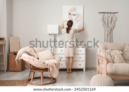 Young pretty woman hanging picture on white wall in stylish living room