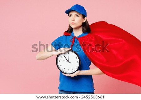 Professional delivery girl employee woman wear blue cap t-shirt uniform workwear red super hero suit work as dealer courier hold in hand clock isolated on plain pastel pink background. Service concept