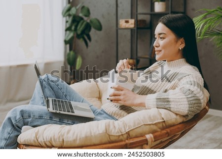 Young IT woman wear casual clothes sits on armchair hold use work on laptop pc computer eat Chinese food stay at home hotel flat rest relax spend spare time in grey living room indoor. Lounge concept