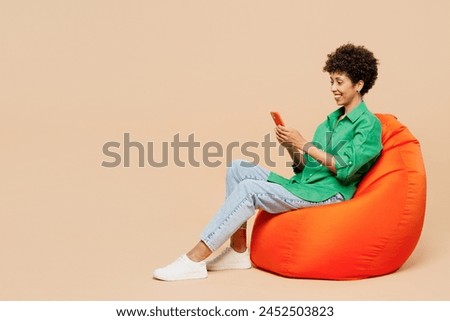 Full body young woman of African American ethnicity wears green shirt casual clothes sit in bag chair hold in hand use mobile cell phone isolated on plain pastel beige background. Lifestyle concept