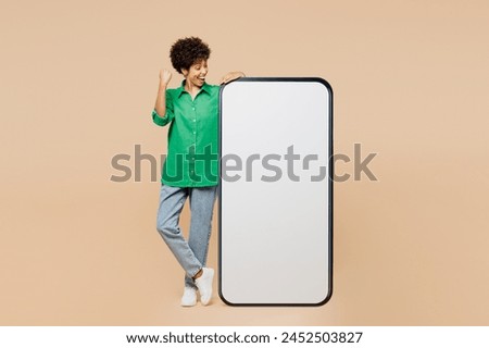 Full body young woman of African American ethnicity wear green shirt casual clothes big huge blank screen mobile cell phone smartphone with area do winner gesture isolated on plain beige background