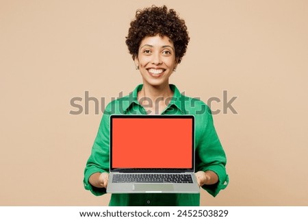 Young IT woman of African American ethnicity wears green shirt casual clothes hold use work on blank screen area laptop pc computer look camera isolated on plain beige background. Lifestyle concept