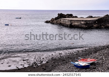 A rocky beach with a few boats on it. The boats are blue and red. The water is calm and the sky is cloudy