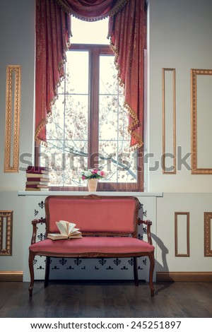 Classic interior design. Elegant interior in red and golden colors with a barocco couch with an open book standing against the window decorated with beautiful curtains and books on the window sill