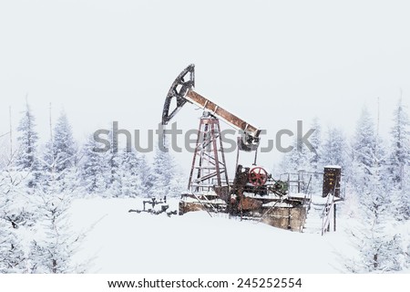 Oil pump. Oil and gas industry equipment. Oil field pump jack and oil refinery in the winter with snow, mountains and forest in background. In a snowy haze.