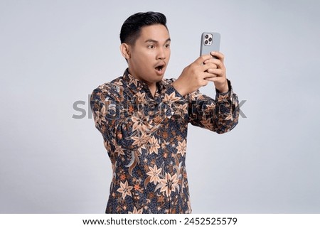Surprised young handsome Asian man wearing batik shirt using smartphone while open mouth iisolated on grey background