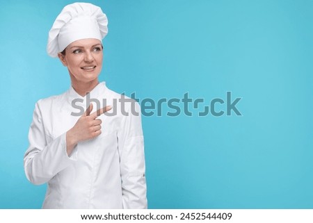 Happy woman chef in uniform pointing at something on light blue background, space for text