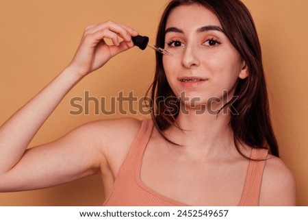 Teenage girl applying cleansing serum against acne and pimples on her cheek. Pretty girl with braces is smiling and holding pipette. Hygiene and skin care in adolescence. Isolated beige background.