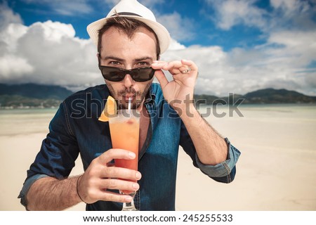 Close up picture of a young casual man on the beach drinking a orange cocktail with a straw while taking off his sunglasses.