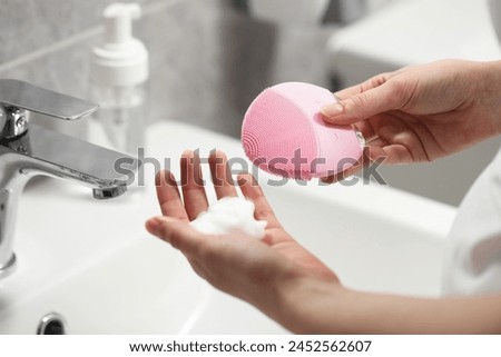 Washing face. Woman with brush and cleansing foam above sink in bathroom, closeup