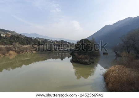 View of the Mtkvari (Kura) river from the bridge in the city of Mtskheta. Shores on both sides, high mountain. Rocky island with trees. Reflection of the sky and trees in the water. Blue sky. Royalty-Free Stock Photo #2452566941