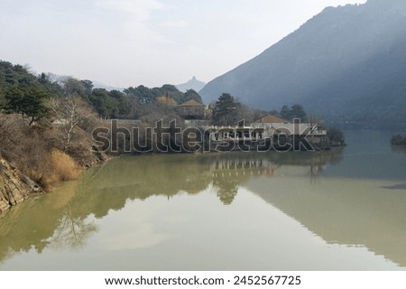 View of the Mtkvari (Kura) river and Jvari monastery from the bridge in the city of Mtskheta. Reflection of the sky, buildings and trees in the water. Royalty-Free Stock Photo #2452567725