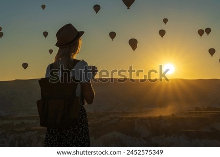 Young female traveler in a hat with a backpack stands alone at the top and admires the flight of balloons during a beautiful sunset, Cappadocia, Turkey. Digital detoxification and soul searching Royalty-Free Stock Photo #2452575349