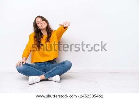 Young caucasian woman sitting on the floor isolated on white wall giving a thumbs up gesture