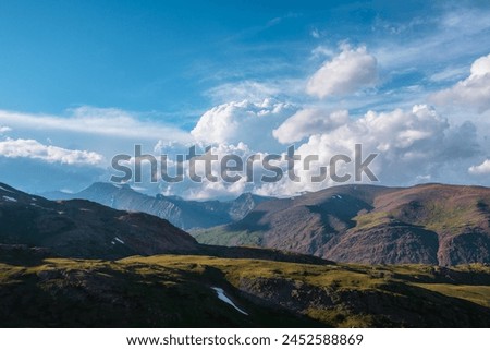 Awesome alpine landscape with sunlit green hills and multicolor mountains under huge lush clouds in blue sky.  Colorful hilly valley and mountain range under dramatic cloudy sky in changeable weather.