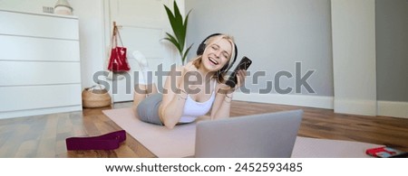 Portrait of woman lying on rubber yoga mat in room, wearing headphones, listens to music, using laptop and smartphone. Lifestyle and wellbeing concept