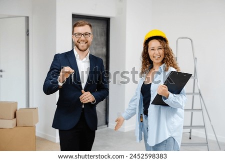 Female designer in helmet with documents giving keys from apartment to young businessman while standing in room.